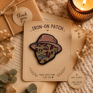 May include: An embroidered iron-on patch featuring a stylised character face with a brown fedora hat, textured skin, and a wide grin. The patch is mounted on a rectangular tan card labelled IRON-ON PATCH, MADE WITH CARE, and EASY TO APPLY.
