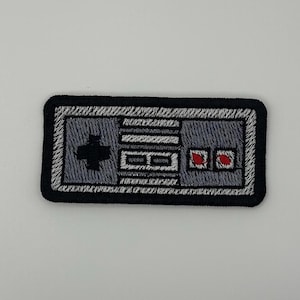 May include: A rectangular embroidered patch featuring a retro video game controller design. The patch has a black border, a grey body, a black directional pad on the left, two small red buttons on the right, and two horizontal grey buttons in the center.