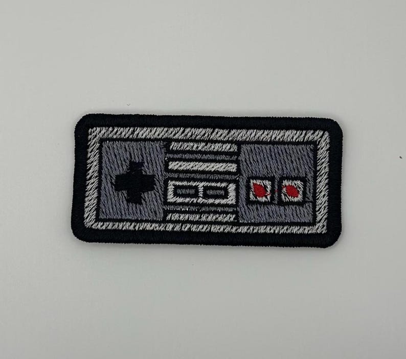 May include: A rectangular embroidered patch featuring a retro video game controller design. The patch has a black border, a grey body, a black directional pad on the left, two small red buttons on the right, and two horizontal grey buttons in the center.