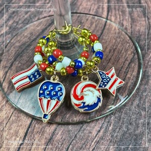 May include: A wine glass charm featuring a circular ring of red, white, and blue beads. Four metal charms hang from the ring: a rectangular flag, a hot air balloon, a circular eagle emblem, and a star, all decorated in red, white, and blue enamel.