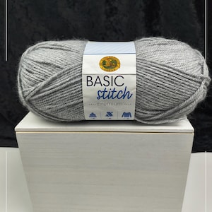 Op de afbeelding: Een bol lichtgrijs garen met een wit papieren label met het Lion Brand Yarns logo en de tekst BASIC stitch premium. Het label bevat kleine iconen van een muts, wanten en een trui. Het garen heeft een zachte, getextureerde uitstraling.