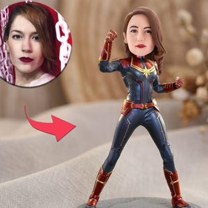 May include: A custom bobblehead figurine featuring a person's likeness with brown hair, wearing a dark blue and red superhero suit with gold accents, including a star emblem on the chest, red gauntlets, and red boots, standing in a dynamic pose.