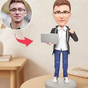 Op de afbeelding: Een op maat gemaakte bobblehead-figuur van een man met een bril, een wit overhemd, een blauwe spijkerbroek en een zwarte jas, die een grijze laptop vasthoudt. Het beeldje staat op een grijze cirkelvormige basis. Een foto van de man staat in een cirkel op de achtergrond.