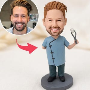 May include: A custom bobblehead figurine depicting a person with brown hair and a beard, wearing a blue tunic with black toggles and dark trousers. The figure holds a pair of grey dental forceps grasping a small white tooth, standing on a base.