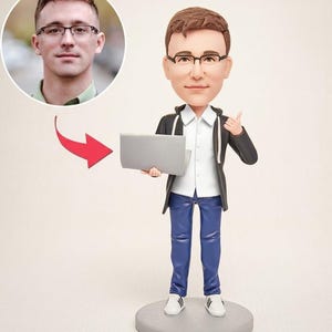 May include: A custom bobblehead figurine depicting a person with brown hair and glasses, wearing a white collared shirt under a dark zip-up hoodie, blue trousers, and white sneakers. The figure holds a grey laptop and stands on a circular base.
