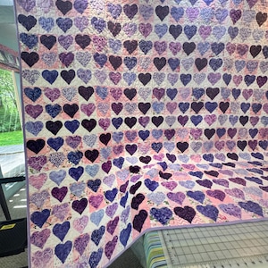 May include: A quilt featuring a repeating pattern of purple and lavender heart-shaped fabric patches arranged in a grid. The quilt has a light purple border and is displayed over a surface with a visible ruler showing measurements in inches.