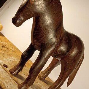 May include: A dark brown wooden horse figurine standing in a profile position. The sculpture features a smooth, polished finish with visible wood grain patterns, pointed ears, a carved mane, and four legs positioned to suggest a standing stance.