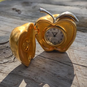 May include: A gold-colored apple-shaped Timex quartz clock with a hinged lid that opens to reveal a white clock face with black numerals and hands. The apple features a silver-colored stem and leaf detail on top, presented next to a small coin.