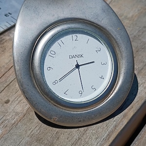 May include: A silver-toned Dansk clock with a white face and black numerals. The clock has a unique, rounded shape and is set on a wooden surface. The clock hands point to approximately 9:28.