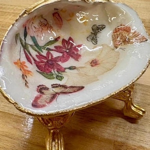 May include: A decorative seashell bowl with a gold-painted rim and base. The interior features a floral and butterfly design in shades of pink, red, and orange. The bowl is elevated on three gold-colored feet.