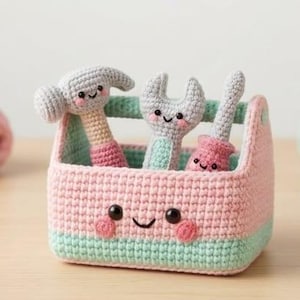 May include: A crocheted tool box in pastel pink and mint green featuring a smiling face with black eyes and pink cheeks. Inside the box are three crocheted tools: a hammer, a wrench, and a screwdriver, each with a smiling face and soft textures.