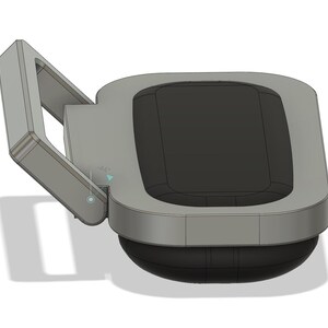 Puede incluir: Un dispositivo gris y negro con una parte superior rectangular y una base curva. El dispositivo tiene un brazo articulado en el lado izquierdo. La superficie superior es negra y los bordes son grises.