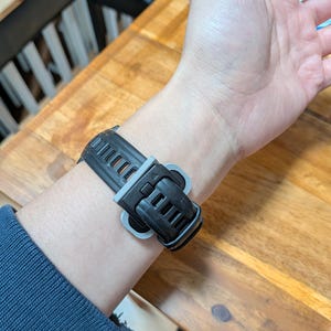 Puede incluir: Una correa de reloj inteligente negra y gris est&aacute; sujeta a una mu&ntilde;eca. La correa est&aacute; hecha de un material flexible con un dise&ntilde;o texturizado. El cierre es negro con un acento gris. La mu&ntilde;eca lleva una camisa azul de manga larga.