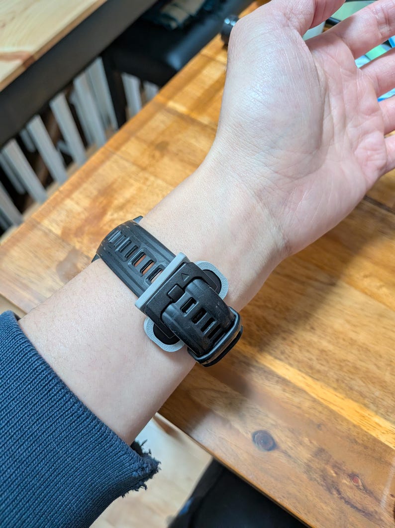 Puede incluir: Una correa de reloj inteligente negra y gris est&aacute; sujeta a una mu&ntilde;eca. La correa est&aacute; hecha de un material flexible con un dise&ntilde;o texturizado. El cierre es negro con un acento gris. La mu&ntilde;eca lleva una camisa azul de manga larga.