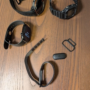 Puede incluir: Un surtido de relojes inteligentes y accesorios, que incluyen un reloj digital negro y naranja, un reloj digital negro y un reloj inteligente gris. Tambi&eacute;n se incluyen correas de reloj y un monitor de frecuencia card&iacute;aca negro.
