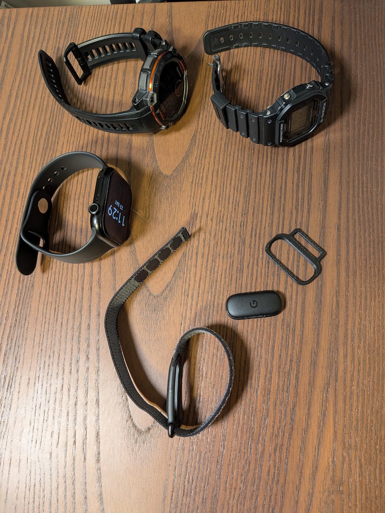 Puede incluir: Un surtido de relojes inteligentes y accesorios, que incluyen un reloj digital negro y naranja, un reloj digital negro y un reloj inteligente gris. Tambi&eacute;n se incluyen correas de reloj y un monitor de frecuencia card&iacute;aca negro.