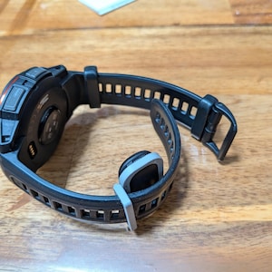 Puede incluir: Reloj inteligente negro con una correa negra texturizada y una hebilla gris. La esfera del reloj tiene detalles en naranja y una parte trasera negra con sensores. La correa est&aacute; abierta, mostrando la parte inferior del reloj.