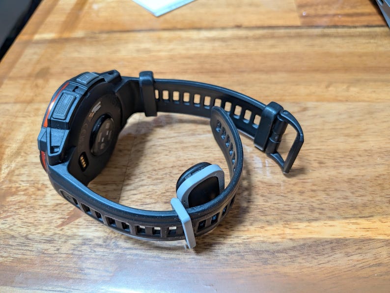 Puede incluir: Reloj inteligente negro con una correa negra texturizada y una hebilla gris. La esfera del reloj tiene detalles en naranja y una parte trasera negra con sensores. La correa est&aacute; abierta, mostrando la parte inferior del reloj.
