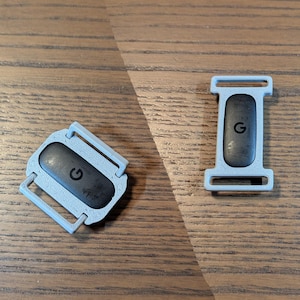 以下が含まれることがあります： ライトグレーのテクスチャ加工されたフレームと滑らかなダークグレーの中央インサートを備えた、2つの長方形の時計バンドコネクタ。各ピースの暗い表面の中央には小さな黒いGのロゴがあり、ストラップを取り付けるためのループが一体化されています。