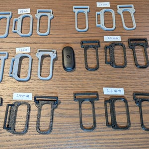 May include: A collection of grey and black plastic strap connectors in various sizes labelled 18mm, 20mm, 21mm, 22mm, 24mm, and 26mm, arranged alongside a small black oval device with a G logo, featuring rectangular loops and side attachment points.