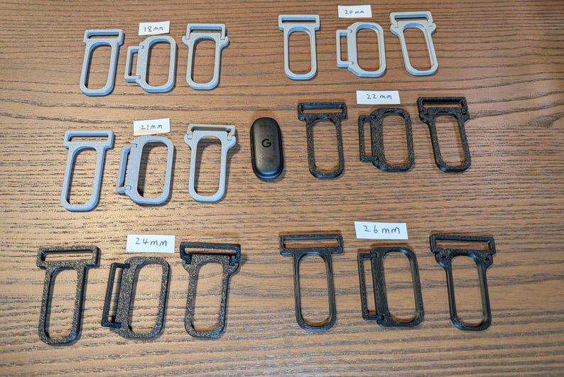 May include: A collection of grey and black plastic strap connectors in various sizes labelled 18mm, 20mm, 21mm, 22mm, 24mm, and 26mm, arranged alongside a small black oval device with a G logo, featuring rectangular loops and side attachment points.