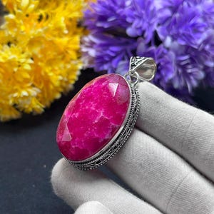 Peut inclure: Un pendentif en pierre précieuse rose vif de forme ovale et à facettes, serti dans une monture en argent décorative avec un travail de métal complexe sur les bords. Le pendentif présente une surface polie et une bélière en argent.