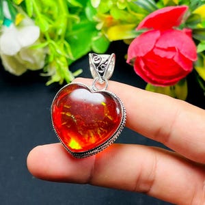May include: Heart-shaped amber pendant with a silver bail. The amber has a warm, reddish-orange hue and is set in a detailed silver frame. The pendant is held up by a finger against a backdrop of flowers.
