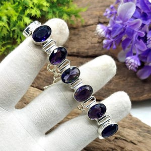 May include: A silver link bracelet featuring seven oval-cut deep purple amethyst gemstones. Each stone is set in a polished silver bezel and connected by silver links, creating a continuous chain design that showcases the dark violet color.