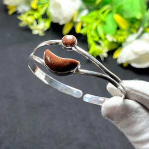 May include: A silver cuff bracelet featuring a crescent moon-shaped brown goldstone and a small round brown goldstone cabochon. The open-ended band is constructed from two thin, polished silver wires that meet at the ends, creating a sleek design.
