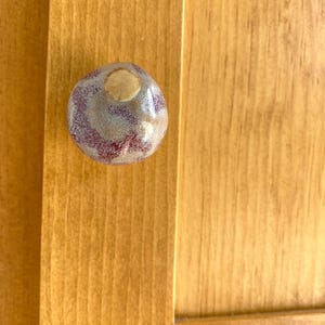 May include: A round, ceramic cabinet knob featuring a glossy, mottled glaze in shades of deep burgundy, soft lavender, and creamy beige. The surface has a smooth, reflective finish with a distinct, circular light-colored patch at the center.