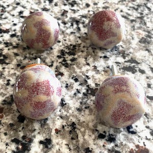 May include: Four round ceramic cabinet knobs featuring a glossy finish with a mottled pattern of dusty rose, cream, and light gray. Each knob has a smooth, spherical shape and includes a metal bolt extending from the back for installation.