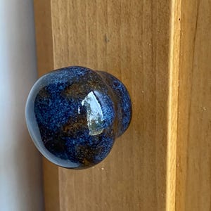 May include: A blue and brown ceramic cabinet knob with a rounded shape. The knob is attached to a light brown wooden cabinet door. The knob has a glossy finish and a speckled pattern.