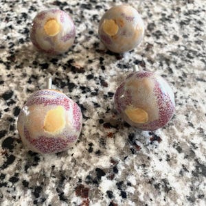 May include: Four round ceramic cabinet knobs with a glossy finish. Each knob features a mottled pattern of light gray, cream, and speckled reddish-purple tones. The knobs have a smooth, rounded shape with a small metal mounting screw protruding from the back.