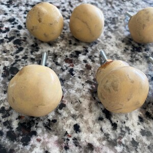May include: A set of five round, light tan cabinet knobs with a smooth, matte finish. Each knob features a central metal bolt extending from the back for installation. The knobs have a natural, slightly mottled appearance with subtle color variations.