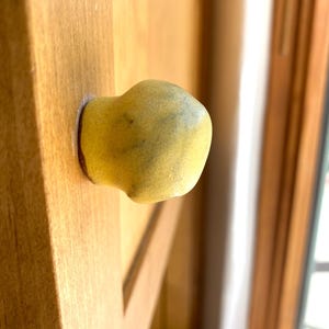 May include: A rounded, mustard-yellow ceramic cabinet knob with a matte, slightly textured surface and subtle dark speckles, attached to a light-toned wooden cabinet door panel with a visible grain pattern and a smooth, finished surface.