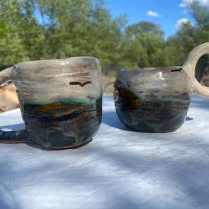 May include: Two handmade ceramic mugs with irregular, organic shapes. The mugs feature a glaze transition from a creamy off-white top to a dark, earthy base with streaks of green and brown. Each mug has a distinct, thick, rounded handle.