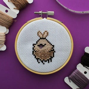 May include: A cross-stitch embroidery hoop showing a cartoon creature stitched with brown and black thread. The creature has a fluffy body, small ears, and a purple mouth. The embroidery is in a wooden hoop, with thread spools visible in the background.