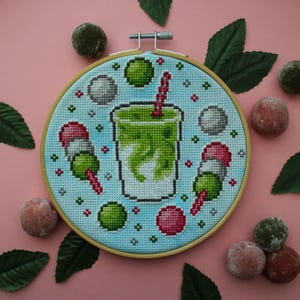 Puede incluir: Un bastidor de bordado circular de madera con un patrón de punto de cruz sobre tela azul claro. El diseño presenta una bebida verde central en un vaso con una pajita a rayas rojas y blancas, flanqueada por dos brochetas dango y esferas.
