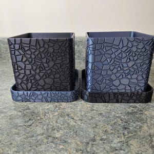 May include: Two square planters with matching trays. One planter is black, the other is a dark blue-gray. Both have a textured, stone-like pattern. The planters are designed for indoor or outdoor use and are suitable for various plants.