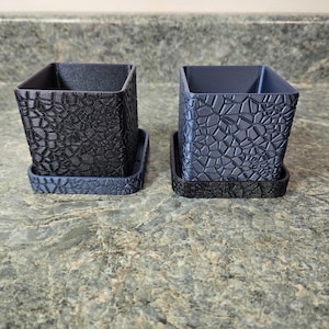 May include: Two square, textured black and gray planters with matching trays. The planters have a geometric pattern and are made of a 3D-printed material. The planters are sitting on a speckled countertop.