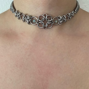 May include: A silver-toned chainmail choker necklace constructed from interlocking metal rings. The design features a central decorative knot-like pattern flanked by two small, iridescent, round beads integrated into the metal link structure.