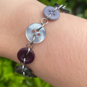May include: A bracelet constructed from a series of circular buttons in varying shades of purple, gray, and black, each connected by small silver-toned metal jump rings. The buttons feature four holes in the center and have a smooth, matte finish.