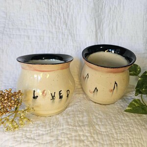 May include: Two ceramic mugs with a cream-colored speckled glaze and a black rim. One mug features the word "LOVED" in black lettering, while the other is decorated with black musical note symbols. Both mugs have a rounded, bulbous shape.
