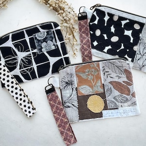 May include: Three rectangular fabric zipper pouches with wrist straps. One features a black and white grid pattern with a floral patch, another has black and white abstract floral shapes, and the third displays a brown and gray botanical collage.