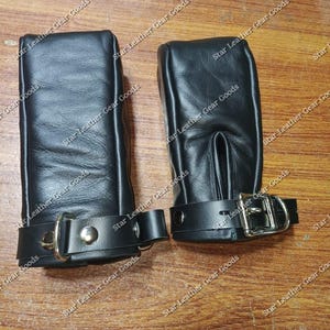 Op de afbeelding: Twee zwarte leren manchetten met een gladde textuur en een rechthoekige vorm. Elke manchet is voorzien van een zwarte leren riem met een zilverkleurige metalen gesp en een D-ring, vastgezet met zilverkleurige klinknagels. De tekst Star Leather Gear Goods is zichtbaar.