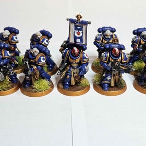Puede incluir: Un grupo de figuras en miniatura de marines espaciales con armadura azul y detalles dorados. Cada figura sostiene un arma y se encuentra sobre una base circular con hierba verde. Una figura sostiene una bandera con un símbolo.