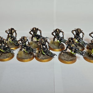 May include: A group of ten miniature robotic figures with metallic bronze bodies and skeletal frames. Each figure holds a black rifle with bright green glowing accents. The figures are mounted on circular bases with textured brown ground.