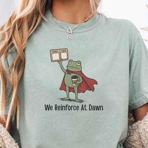 May include: A light green t-shirt featuring a cartoon frog superhero holding a coffee cup and a sign that says "First Then". The frog wears a red cape and the text "We Reinforce At Dawn" is printed below the frog.