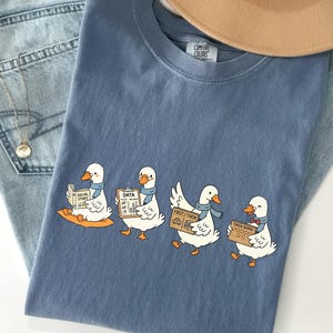 Pode incluir: Uma camiseta azul Comfort Colors com um gráfico de quatro patos de desenho animado, cada um segurando uma placa com texto. Os patos estão usando cachecóis. A camisa está sobre jeans azul claro, um colar de ouro e um chapéu bege.