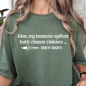 Peut inclure: T-shirt vert olive avec le texte blanc "Alas, my immune system hath chosen violence... once more" et un dessin de poignard. Le t-shirt est de style décontracté.